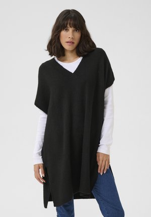 Poncho noir en tricot avec un col en V, des manches courtes et des fentes sur les côtés, superposé sur une chemise blanche à manches longues. Texture douce, coupe ample.