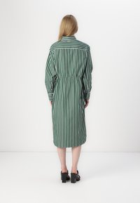 Rochie verde în dungi cu mâneci lungi, talie strânsă și lungime sub genunchi. Prezintă un guler clasic și manșete cu nasturi. Pantofi cu toc pătrat negri.