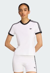 Biele bavlnené tričko s čiernymi akcentmi, krátkymi rukávmi a okrúhlym výstrihom; na ramenách sú tri čierne pruhy a logo Adidas.