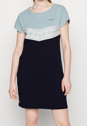 Nainen pukeutuneena lyhythihaiseen mekkoon, jonka yläosa on sininen, keskellä valkoinen kuvioitu vyö ja alaosa tummansininen. Hänellä on helmikaulakoru.