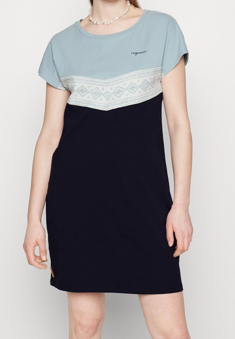 Femme portant une robe à manches courtes avec une partie supérieure bleue, une bande blanche à motifs et un bas bleu marine foncé, accessoirisée d'un collier de perles.