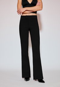 Fekete, bővülő szárú nadrág sima anyagból, testhezálló derékkal, letisztult elülső résszel, hegyes orrú fekete cipővel párosítva.