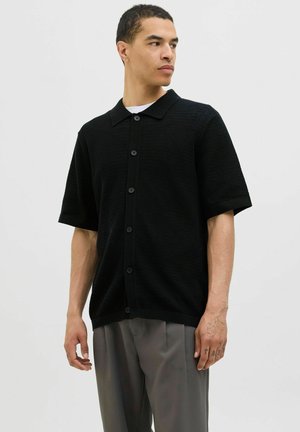 Black knit short-sleeve shirt with a polo collar, button-down front, and textured fabric. Worn with gray trousers, featuring a relaxed fit.