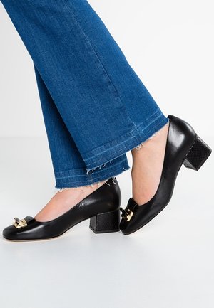 Sorte lædersko med lav blokhæl og guldlåsdetalje, båret med blå jeans med frynset kant på krydsede ben.