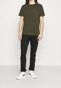 Oliverová zelená trička s kapsou na hrudi a zipem, spárovaná s černými slim-fit džíny a bílými teniskami s černými akcenty.