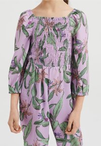 Lila Jumpsuit mit gerafftem Oberteil, langen Puffärmeln und einem floralen Muster mit grünen Blättern und verschiedenen bunten Blumen.
