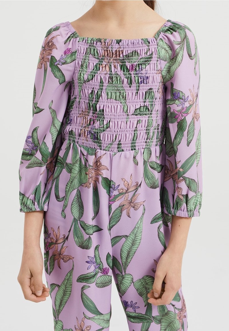 Lila Jumpsuit mit gerafftem Oberteil, langen Puffärmeln und einem floralen Muster mit grünen Blättern und verschiedenen bunten Blumen.