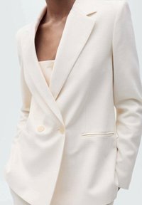 Blazer sur mesure couleur crème avec un design à double boutonnage, des revers crantés et une texture subtile. Comprend deux poches avant et des boutons clairs.