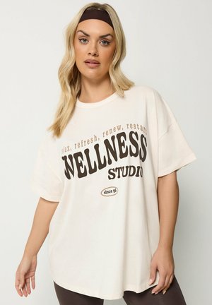 Blondine draagt een wit oversized T-shirt met de tekst "relax, refresh, renew, restart WELLNESS STUDIO since 96" in bruine letters, zwarte hoofdband.
