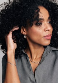 Srebrne kolczyki w kształcie obręczy, falista srebrna naszyjnik i bransoletka noszone przez osobę z kręconymi włosami, w jasnoszarej koszuli zapinanej na guziki.