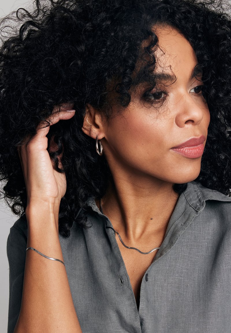 Srebrne kolczyki w kształcie obręczy, falista srebrna naszyjnik i bransoletka noszone przez osobę z kręconymi włosami, w jasnoszarej koszuli zapinanej na guziki.