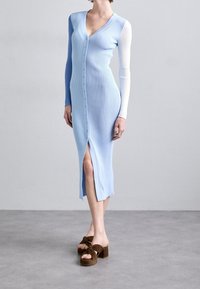 Žena oblečená v dlouhých, ribbovaných, propínacích modrých šatech s jedním bílým rukávem a hnědými podpatkovými sandály, stojící na šedé podlaze.