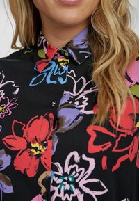 Camicia a maniche corte con bottoni, caratterizzata da uno sfondo nero con un vivace motivo floreale multicolore e colletto. Sua trama di tessuto liscia.