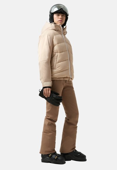 Beżowa kurtka puchowa o pikowanej fakturze, brązowe spodnie narciarskie, czarne rękawice i buty narciarskie. Dodatki to czarny kask z odblaskową wizjerem.