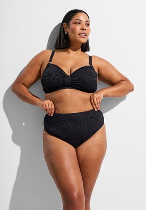 Žena s čiernymi vlasmi po ramená, ktorá má na sebe čierne vzorované dvojdielne plavky, pózuje pred bielou stenou odrážajúcou tieň.