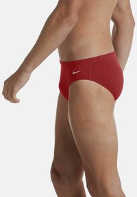 Slip da nuoto rossi con una texture liscia, caratterizzati da un logo Nike bianco sul lato. Il design presenta un taglio a vita bassa con copertura minima.