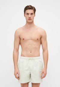 Maillots de bain vert clair avec taille à cordon de serrage et logo brodé sur le côté. Le matériau est lisse, adapté aux activités aquatiques.