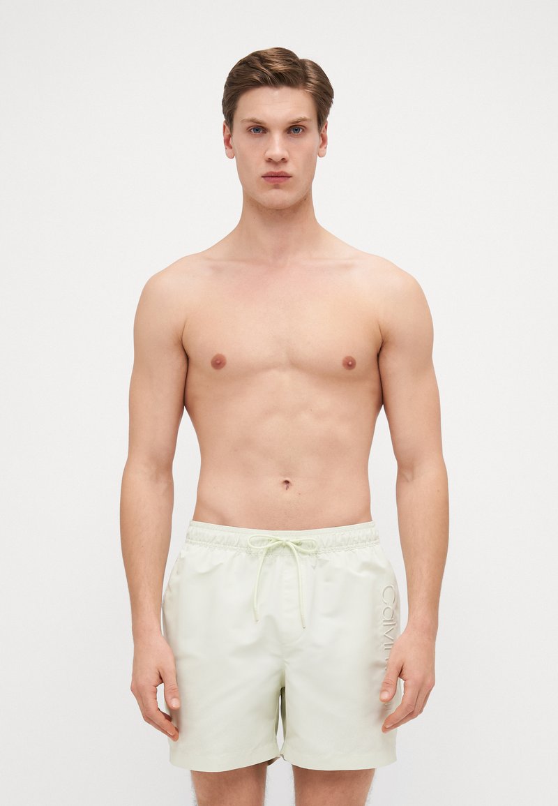 Maillots de bain vert clair avec taille à cordon de serrage et logo brodé sur le côté. Le matériau est lisse, adapté aux activités aquatiques.