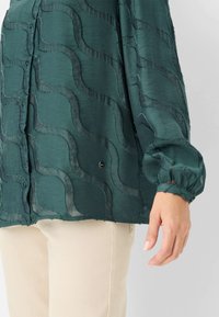 Green blouse in lightweight fabric with wavy texture, buttoned front, and elastic cuffs. Smooth finish with subtle sheen, paired with beige pants.