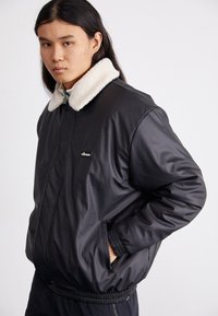 Chaqueta bomber negra con un collar de piel beige, frontal con cremallera y puños elásticos. Presenta un pequeño logo en el pecho y bolsillos laterales.