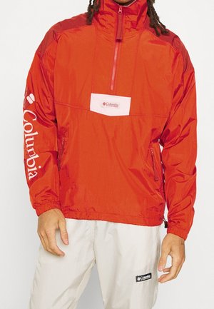 Person trägt eine rote Columbia Quarter-Zip-Jacke mit Logo auf Brust und Ärmel, kombiniert mit hellbeigen Hosen mit Columbia Logo-Patch.