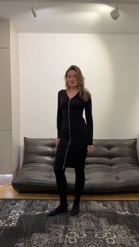 Woman with long hair wearing a black long-sleeve midi dress with front zipper stands barefoot on a patterned rug in a modern living room.