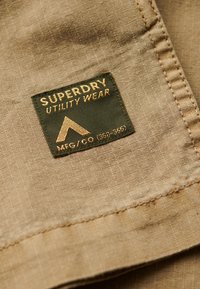 Étiquette en tissu beige avec "SUPERDRY UTILITY WEAR" en texte doré et un fond vert, incluant un logo triangulaire et des détails de fabrication.