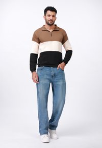 Pull à fermeture éclair marron, crème et noir avec un design coloré, associé à un jean en denim bleu clair et des baskets blanches.
