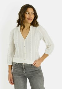 Mujer con cabello ondulado que lleva una blusa blanca con botones y estampado, y jeans grises, con una mano en el bolsillo, mirando hacia un lado y sonriendo.