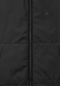 Chaqueta negra vista frontal con cremallera central y pequeño detalle de "X" cosido en la parte superior derecha.