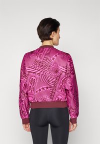 Roze bomberjack met abstracte patronen, featuring een bordeaux ribbelkraag en manchetten. Gladde textuur, ontspannen pasvorm. Zwarte leggings zichtbaar eronder.