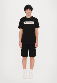 Svart bomull t-skjorte med korte ermer, med et kremfarget merke som har "A|X ARMANI EXCHANGE." Kombinert med svarte shorts og hvite sneakers.
