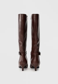 Par de botas altas de cuero marrón oscuro con tacones bajos y correas en el tobillo, vistas desde atrás contra un fondo claro y liso.