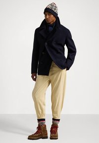 Cappotto doppiopetto in lana blu navy, pantaloni in velluto beige, cappello a maglia con motivo, calzini lavorati a maglia e stivali da trekking marroni con lacci rossi.