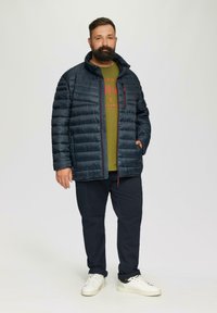 Marineblaue Steppjacke mit horizontalen Nähten, ein Reißverschluss-Kragen, getragen über einem olivgrünen Hemd. Dunkle Hosen und weiße Turnschuhe runden den Look ab.