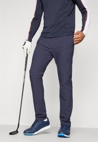 Marine golfbroeken van lichtgewicht materiaal, met een getailleerde pasvorm en een slank ontwerp, gedragen met blauwe gymschoenen. Handschoenen voor grip op een golfclub.