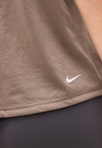 Καφέ ύφασμα με λευκό λογότυπο Nike κοντά στο στριφτό, φορεμένο πάνω από μαύρα κολάν, με λεπτομέρειες ραφής στην κάτω άκρη.