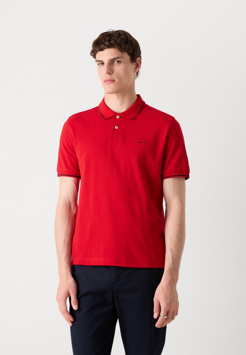 Polo rouge avec un col classique, deux boutons et des accents navy sur le col et les manches. Présente un petit logo sur la poitrine gauche.