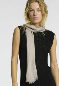 Lys beige tørklæde lavet af blødt stof, draperet afslappet om halsen. Båret over en tekstureret sort ærmeløs top med en strikket mønster.