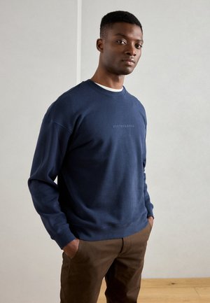 Sweatshirt bleu marine à col rond en tissu doux, avec des poignets et un ourlet côtelés, portant une petite marque ton sur ton sur la poitrine. Pantalon marron.