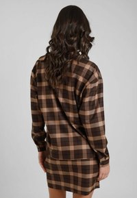 Donna con capelli lunghi e mossi che indossa una camicia a maniche lunghe a quadri marroni e beige e una gonna abbinata, di spalle su uno sfondo semplice.