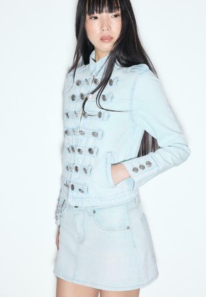 Mladá žena s dlhými čiernymi vlasmi, oblečená v svetlomodrej denimovej bunde so striebornými gombíkmi a zodpovedajúcou denimovou sukňou, stojaca na bielom pozadí.