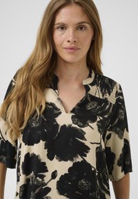 Moteris su ilga šviesiai ruda šukuosena, vilkinti bežė ir juodos spalvos gėlių rašo marškiniais su iškirpte, prieš vientisą foną.