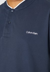 Marineblauwe polo shirt gemaakt van gestructureerde stof; heeft een witte kraag, knoopsluiting en geborduurd 'Calvin Klein' logo op de borst.