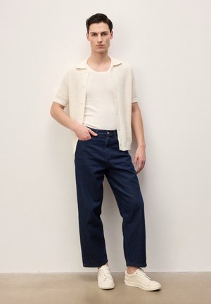 Mladý muž stojí opřený o zeď, má na sobě bílý krátký pletený rukáv přes bílé tílko, tmavě modré džíny a bílé tenisky.