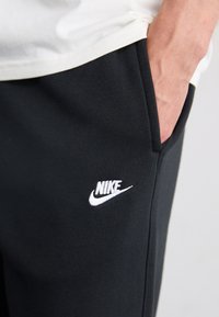 Črne bombažne Nike trenirke imajo belo vezeno logotip na levi stegnici in stransko žep. Tkanina je videti gladka in elastična.