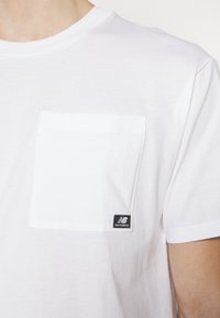 White cotton t-shirt with a crew neck, featuring a square pocket and a small black "New Balance" logo tag on the hem.