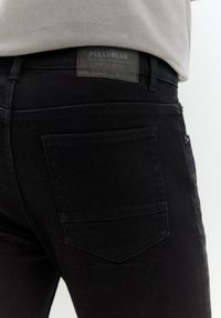 Crne traperice od denima s detaljima na stražnjem džepu, s kožnom oznakom Pull&Bear na pojasu. Jednostavan dizajn, bez ukrasa.