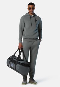 Felpa grigia con cappuccio regolabile, pantaloni della tuta abbinati e borsone grigio. Il modello indossa occhiali da sole amber e sneakers grigie con suole chiare.