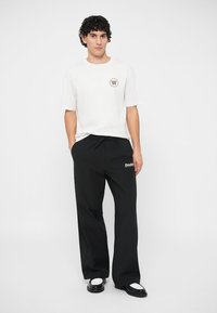 Witte katoenen t-shirt met een bruine logo, gecombineerd met zwart ontspannen pasvorm broeken met een witte merknaam. Schoenen zijn zwart en wit.